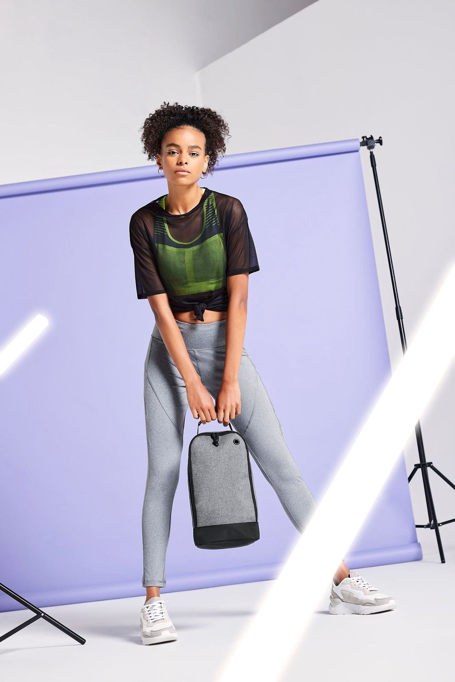 A woman wearing a green top and gray leggings stands in front of a purple backdrop, holding a black bag.
