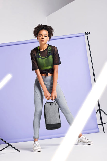 A woman wearing a green top and gray leggings stands in front of a purple backdrop, holding a black bag.